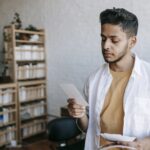 Serious young ethnic male in casual clothes looking attentively at images in hands while standing in modern light living room