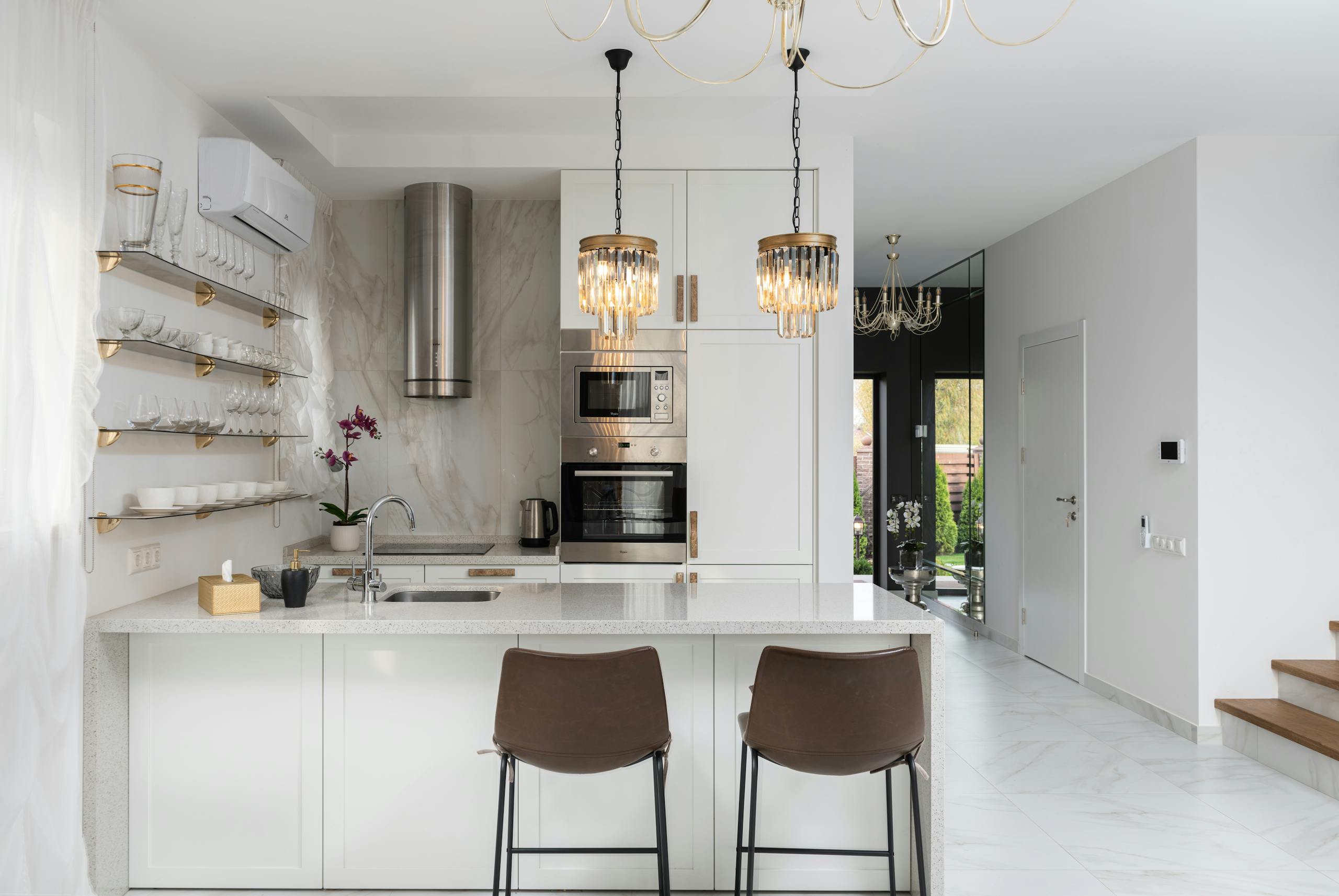 Interior of contemporary kitchen with modern appliances and chairs at white counter near wall with shelves in spacious apartment near stairs